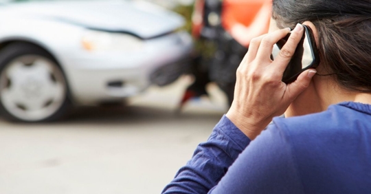 woman with a cell phone to her ear and a car accident scene in front of her.